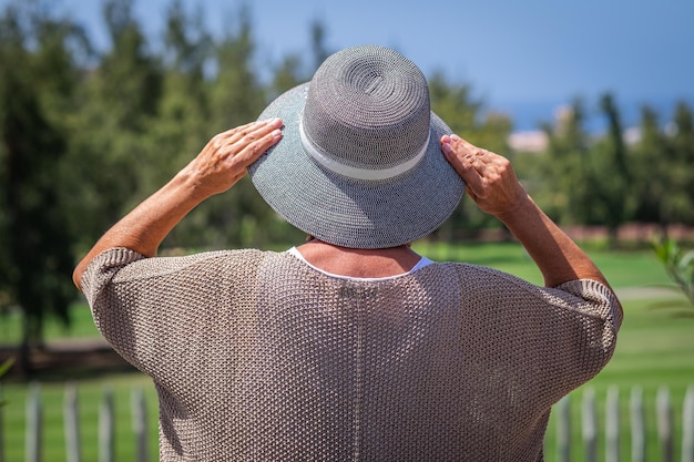 Rückansicht einer reifen kaukasischen Frau, die im öffentlichen Park steht und seinen Hut hält und auf den Horizont blickt