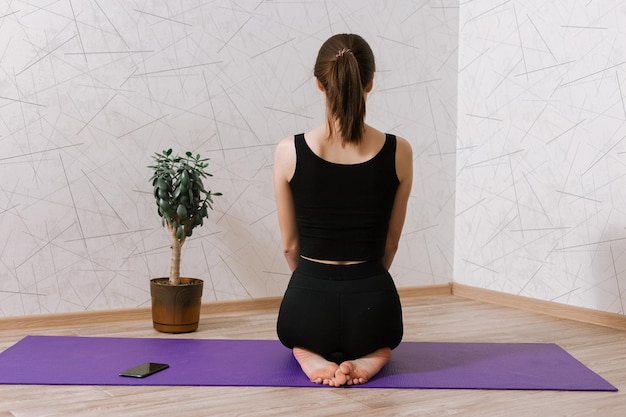 Rückansicht einer schlanken Frau, die auf einer Yogamatte sitzt und während des Trainings zu Hause meditiert