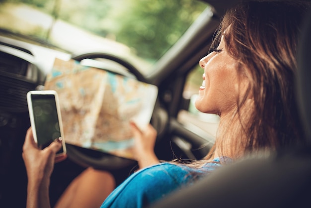 Rückansicht einer schönen jungen lächelnden Frau in einem Auto, die auf die Straßenkarte schaut und ein Smartphone verwendet.