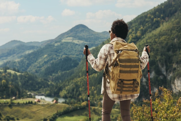 Rückansicht einer schwarzen Frau mit Rucksack und Trekkingstöcken, die in der Natur stehen und beim Wandern auf dem Berg an der frischen Luft genießen.