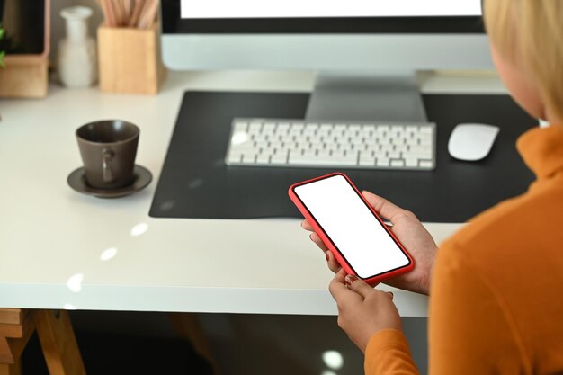 Rückansicht einer stilvollen Frau, die ein Smartphone hält, während sie in einem modernen Büro vor dem Computer sitzt