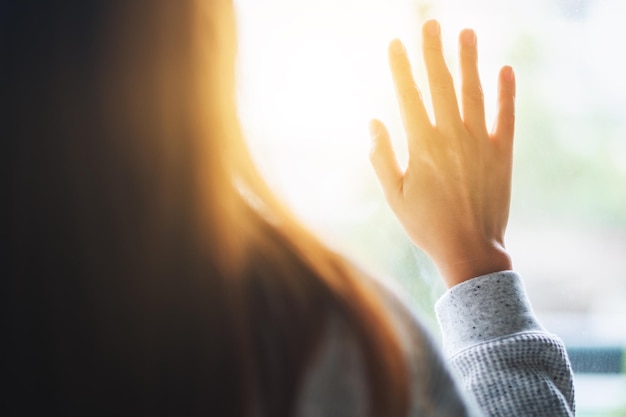 Rückansicht einer traurigen Frau, die ein Fenster berührt, während sie nach draußen schaut