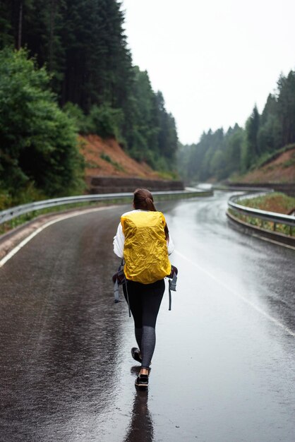 Rückansicht einer Wanderin mit gelbem Rucksack, die auf einer asphaltierten Straße mit wunderschönen Bergen rundherum läuft. Konzept von Menschen und Freizeitaktivitäten