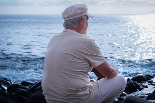 Rückansicht eines älteren kaukasischen Mannes, der am Kiesstrand sitzt und sich entspannt und den Horizont über dem Wasser betrachtet Älterer weißhaariger Mann mit Mütze, der Urlaub oder Ruhestand genießt