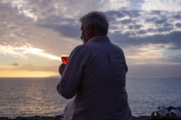 Rückansicht eines älteren Mannes, der bei Sonnenuntergang vor dem Meer sitzt und ein Glas Wein trinkt