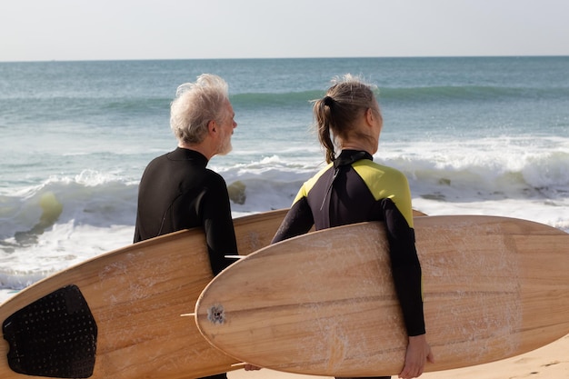 Rückansicht eines älteren Paares mit großen Surfbrettern. Grauhaariger Mann und Frau in Neoprenanzügen, die an der Küste entlang gehen, bevor sie mit dem Surfen beginnen. Sportliche Aktivität und gesunder Lebensstil des Alterskonzepts
