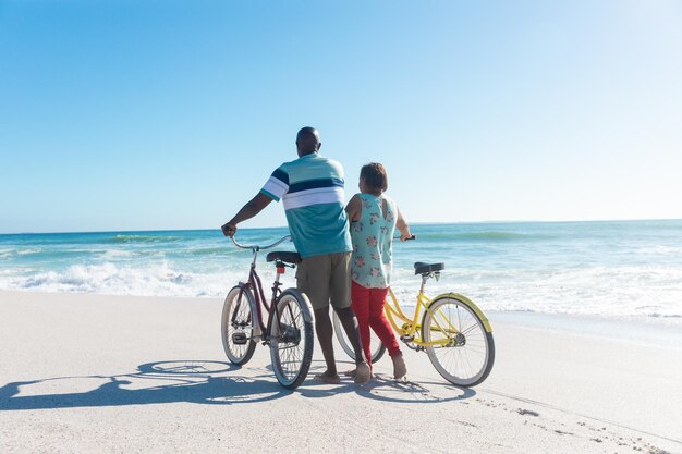 Rückansicht eines afrikanisch-amerikanischen Seniorenpaares, das am Strand Fahrräder fährt, mit Kopierraum am blauen Himmel