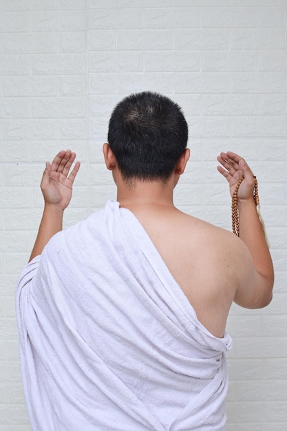 Rückansicht eines asiatischen muslimischen Mannes, der weiße ihram-Kleidung trägt und betet, während er Gebetsperlen hält