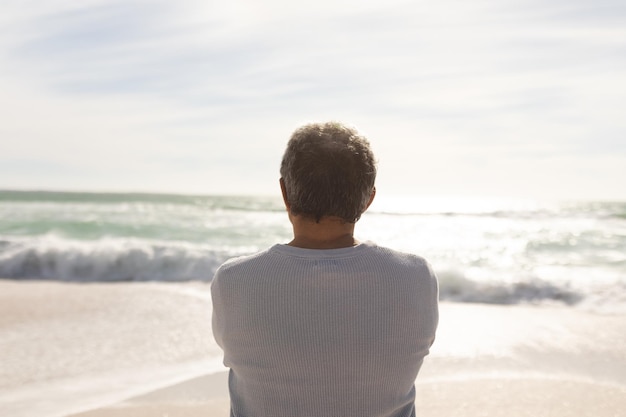 Rückansicht eines biracial älteren Mannes, der vom sonnigen Strand aus auf Wellen blickt, die im Meer gegen den Himmel schlagen