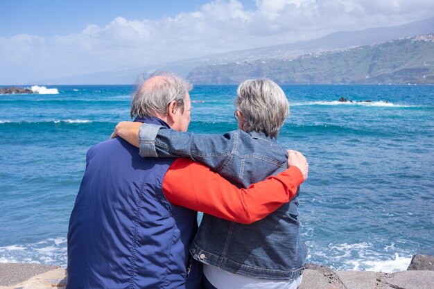 Rückansicht eines entspannten kaukasischen Seniorenpaares, das sich am Strand ausruht und den Horizont über Wasser betrachtet und Urlaub oder Ruhestand genießt