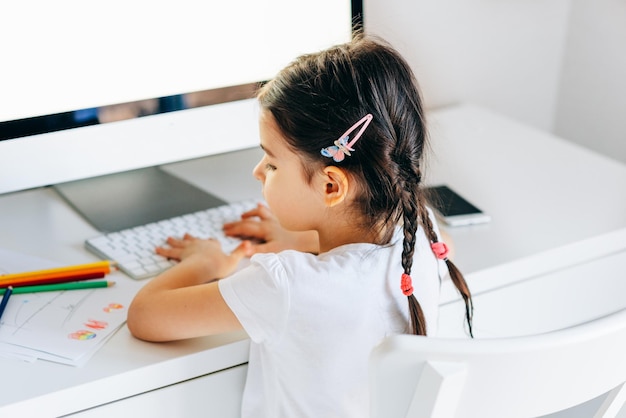 Rückansicht eines entzückenden kleinen Mädchens, das zu Hause mit Bleistiften neben dem Computer zeichnet Kreatives Kind, das an einem Tisch im Zimmer sitzt und etwas auf dem PC eingibt Kleinkindermädchen, das zu Hause Hausaufgaben macht