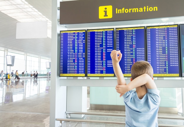 Rückansicht eines gelangweilten Kindes, das seine Arme verschränkt und die Flugzeiten auf dem Flughafen sieht