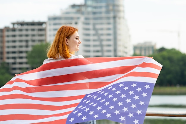 Rückansicht eines glücklichen rothaarigen Mädchens mit USA-Flagge auf ihren Schultern. Positive junge Frau, die den Unabhängigkeitstag der Vereinigten Staaten feiert. Internationaler Tag des Demokratiekonzepts.
