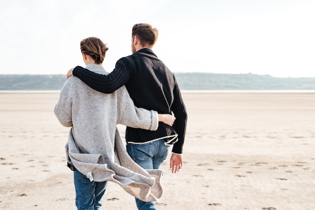 Rückansicht eines jungen, beiläufigen Paares, das sich umarmt und am Strand entlang geht?