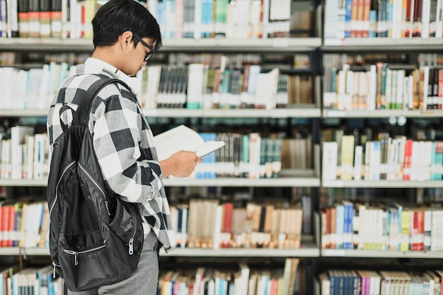 Rückansicht eines jungen männlichen Studenten, der vor den Bücherregalen der Bibliothek ein Buch liest