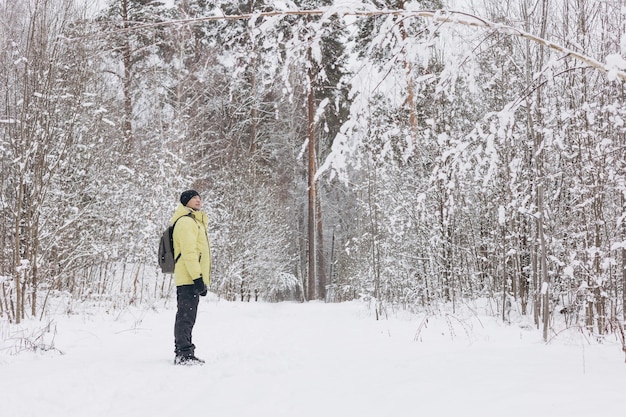 Rückansicht eines jungen Mannes mit Rucksack in gelber Jacke, der im Winter im verschneiten Kiefernwald spazieren geht. Menschen von hinten. Reisen vor Ort, Erkundung der Natur.