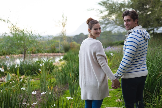 Rückansicht eines kaukasischen Paares, das Händchen haltend durch den Garten geht, sich umdreht und in die Kamera lächelt. Wochenendaktivitäten zu Hause und im Lifestyle.
