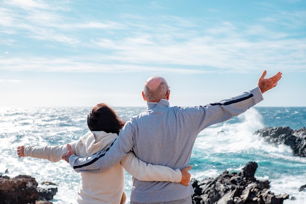 Rückansicht eines kaukasischen, reifen älteren Ehepaares, das mit offenen Armen auf den Horizont über dem Meer blickt