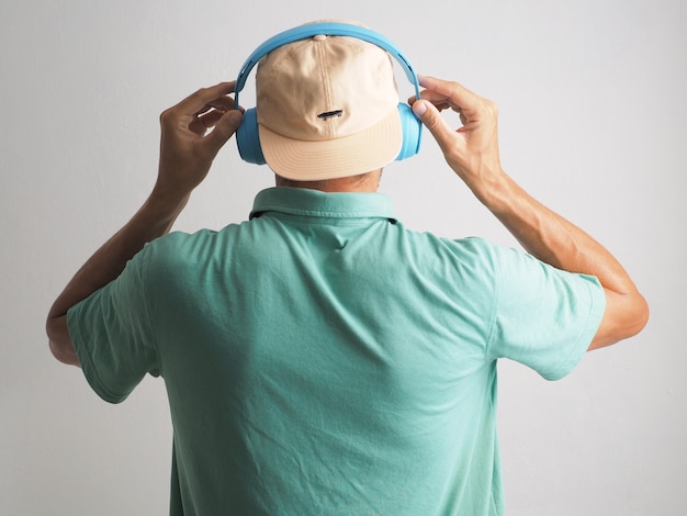 Foto rückansicht eines mannes mit blauen drahtlosen kopfhörern und mütze, der musik gegen die weiße wand hört.