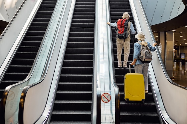 Rückansicht eines Mannes und einer Frau, die die Rolltreppe am Flughafenterminal hinauffahren