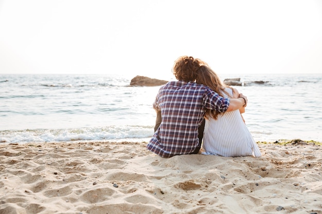 Rückansicht eines schönen jungen Paares, das am Strand sitzt und umarmt