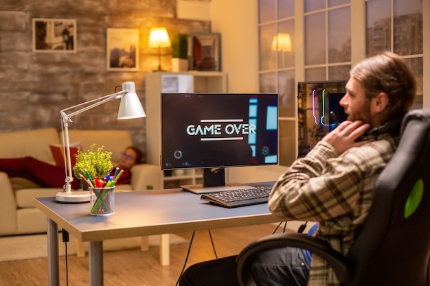 Rückansicht eines Spielers, der spät in der Nacht im Wohnzimmer einen Shooter auf seinem leistungsstarken PC-Computer spielt.