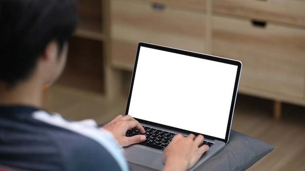 Rückansicht eines zufälligen jungen Mannes, der Computer-Laptop verwendet, während er auf der Couch im Wohnzimmer sitzt.