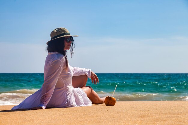 Rückansicht erwachsene Frau in weißem Beachwear-Hut und Sonnenbrille mit Kokosnuss, die auf das Meer blickt