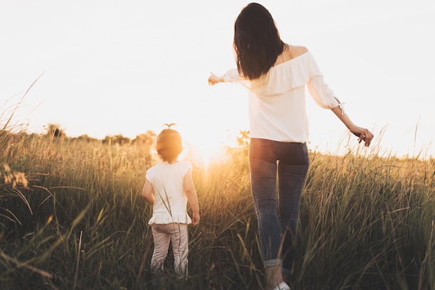 Rückansicht Ganzkörper der kaukasischen Brünette Mutter und Kind Tochter suchen auf Wiese Sonnenuntergang Hintergrund Schöne Frau zeigt ihr Kleinkind Mädchen Wiese Sonnenuntergang Adorable Hintergrund Sonnenuntergang