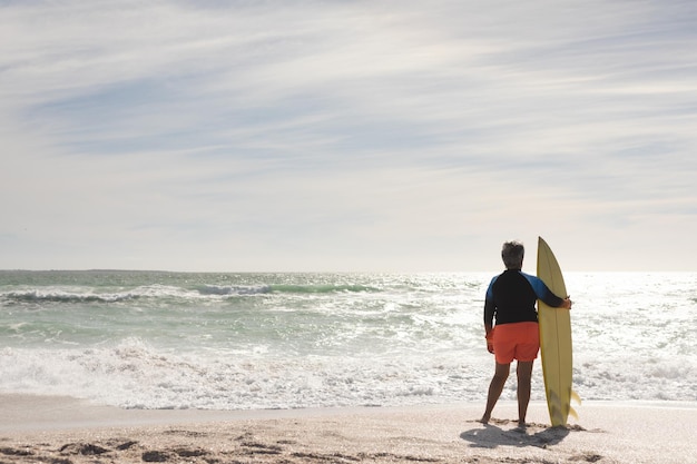 Rückansicht in voller Länge einer älteren biracial Frau mit Surfbrett, die vom sonnigen Strand aus auf den Horizont blickt