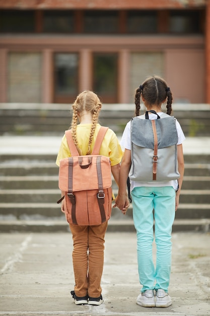 Rückansicht in voller Länge Porträt von zwei niedlichen Schulmädchen mit Rucksäcken, die draußen gegen Schulgebäude stehen und Hände halten