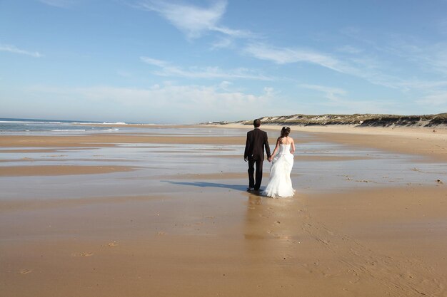 Rückansicht Jungvermählten, die einen romantischen Moment am Strand teilen, Bräutigam und Braut, die am Sandstrand spazieren gehen