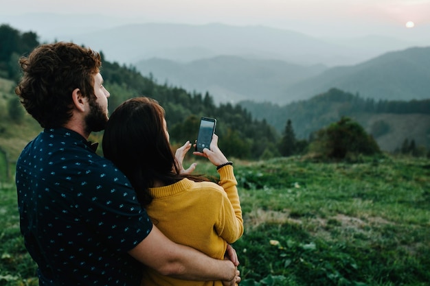 Rückansicht Liebevolles Paar Mann umarmt Frau und genießt die schöne Landschaft auf Tal, Berge und Wälder Summer Mountain auf dem Hintergrund Mädchen fotografiert am Telefon Karpaten Ukraine