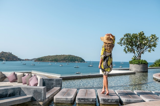 Rückansicht: Luxuriöse Blondine posiert in der Nähe des Infinity-Pools mit herrlichem Blick auf das Meer und die Yachten.