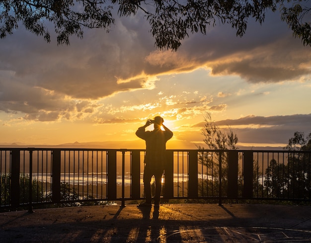 Rückansicht Mann Silhouette Fotografieren mit Telefon in einem Lookout bei Sonnenuntergang in Noosa, Australien