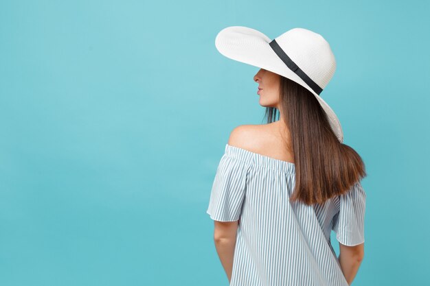 Rückansicht Porträt der eleganten Mode junge Frau im weißen Sommer Sonnenhut mit großer breiter Krempe, Kleid legte die Hände auf den Kopf und schaute auf den Kopienraum einzeln auf blauem Pastellhintergrund. Lifestyle-Konzept.