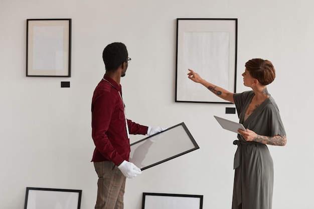 Rückansicht Porträt des weiblichen Kunstgaleriemanagers, der Arbeiter anweist, Malrahmen an weiße Wand zu hängen, während Ausstellung im Museum plant,