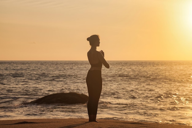 Rückansicht Silhouette schlanke Frau macht Yoga an der tropischen Küste oder am Meeresstrand im Freien bei Sonnenuntergang