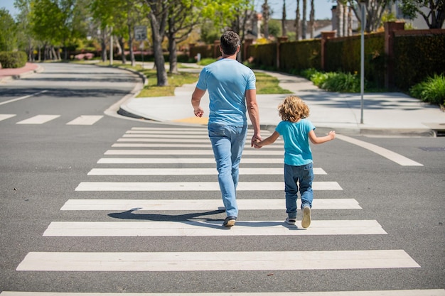 Rückansicht. Vater und Sohn gehen auf Zebrastreifen. Familienwert. Eltern, die kleinen Jungen auf Fußgängerüberweg führen. Familie von Papa mit Kind Kreuzung. Elternschaft und Vaterschaft.