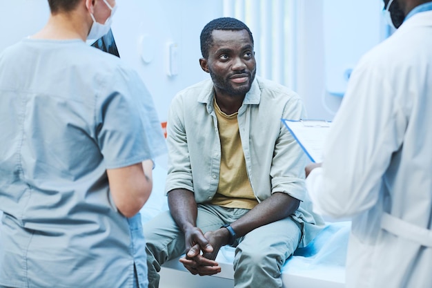 Rückansicht von Ärzten mit Testergebnissen, die mit einem afroamerikanischen Patienten sprechen, der auf einem Untersuchungstisch in der Klinik sitzt