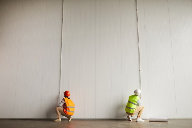 Rückansicht von zwei Arbeitern in reflektierender Kleidung, die mit Drähten an der weißen Wand im leeren Gebäude auf der Baustelle arbeiten