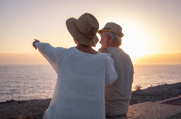 Rückansicht von zwei entspannten und romantischen Senioren oder Rentnern, die bei Sonnenuntergang mit Blick auf das Meer sitzen und auf den Horizont blicken Altes Ehepaar im Freien, das gemeinsam Urlaub macht