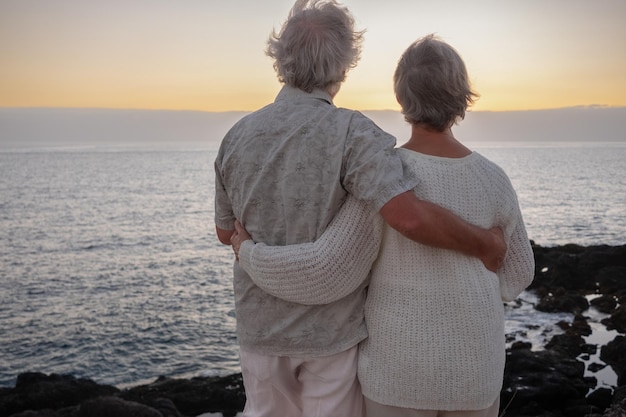 Rückansicht von zwei entspannten und romantischen Senioren oder Rentnern, die sich bei Sonnenuntergang am Meer umarmen und auf den Horizont schauen Altes Ehepaar im Freien, das gemeinsam Urlaub macht