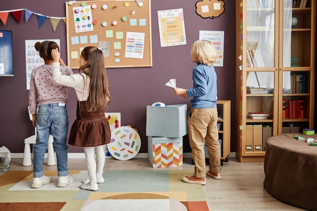 Rückansicht von zwei interkulturellen kleinen Mädchen in schicker Freizeitkleidung und süßem blonden Jungen mit Papierflieger