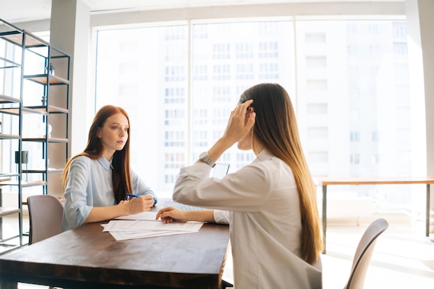 Rückansicht von zwei weiblichen Büroangestellten, die am Schreibtisch mit Papierdokumenten sitzen, die einander anschauen, besprechen Projekt-Brainstorming, das zusammenarbeitet Junge Frau, die ein Vorstellungsgespräch für eine Stelle hat