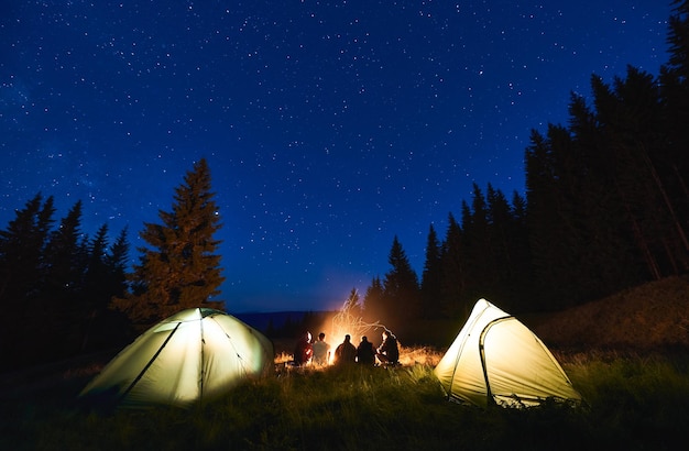 Rückblicktouristen sitzen am hell lodernden Lagerfeuer zwischen den Zelten unter blauem Nachthimmel mit hellen Sternen