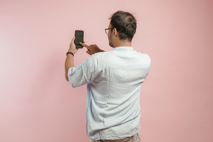 Rückenporträt eines jungen asiatischen Mannes in blauem Hemd, der ein Smartphone hält und benutzt