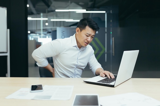 Rückenschmerzen am Arbeitsplatz Junger asiatischer Geschäftsmann, der seinen Rücken am Schreibtisch hält
