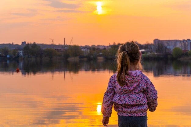 Rückseite des kleinen Mädchens und des Sonnenunterganghimmels in der Stadtreflexion im Fluss