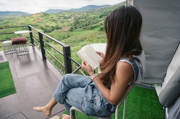 Rückseite einer asiatischen Frau, die auf der Terrasse inmitten der Natur im Urlaub sitzt und ein Buch liest